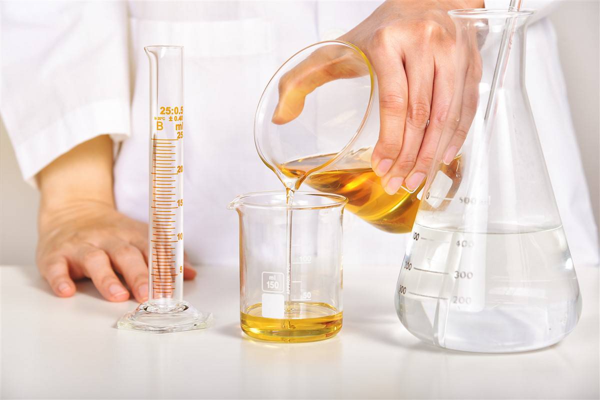 Person in a white lab coat pouring oil from one beaker into another